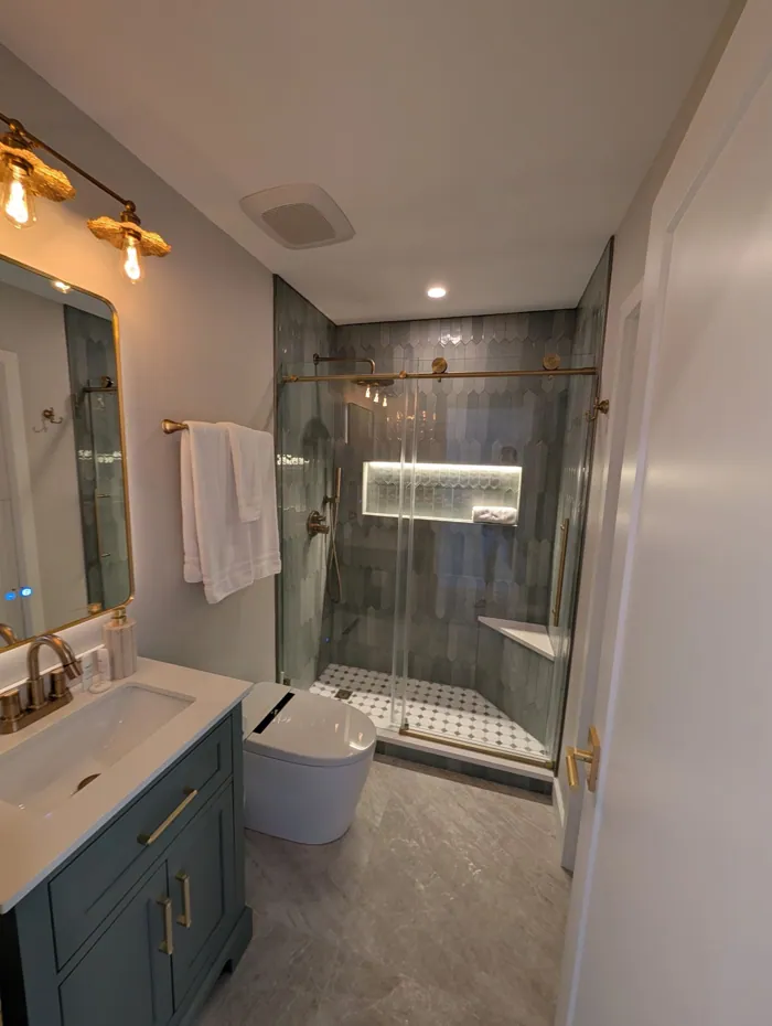 Bathroom with grey tiled walk-in shower, brass fixtures, gold sconces and backlit mirror.