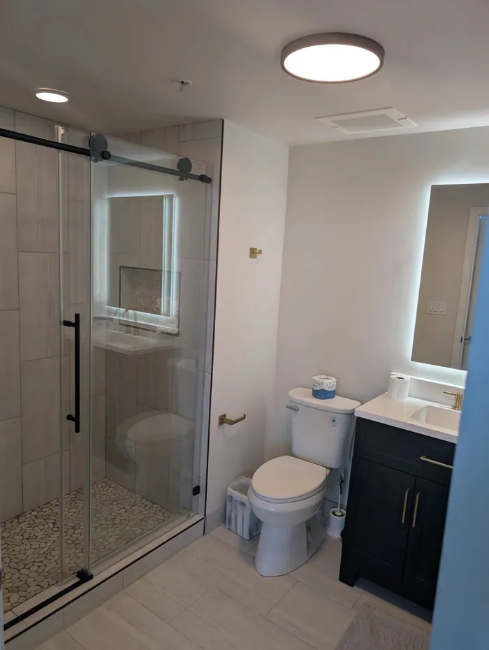 Bathroom with sliding glass shower door, navy vanity, brass pulls and backlit mirror.