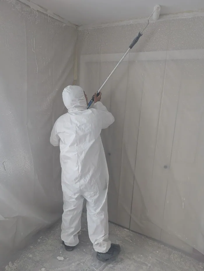 Painter in white protective coveralls spraying inside sealed plastic containment sheeting.